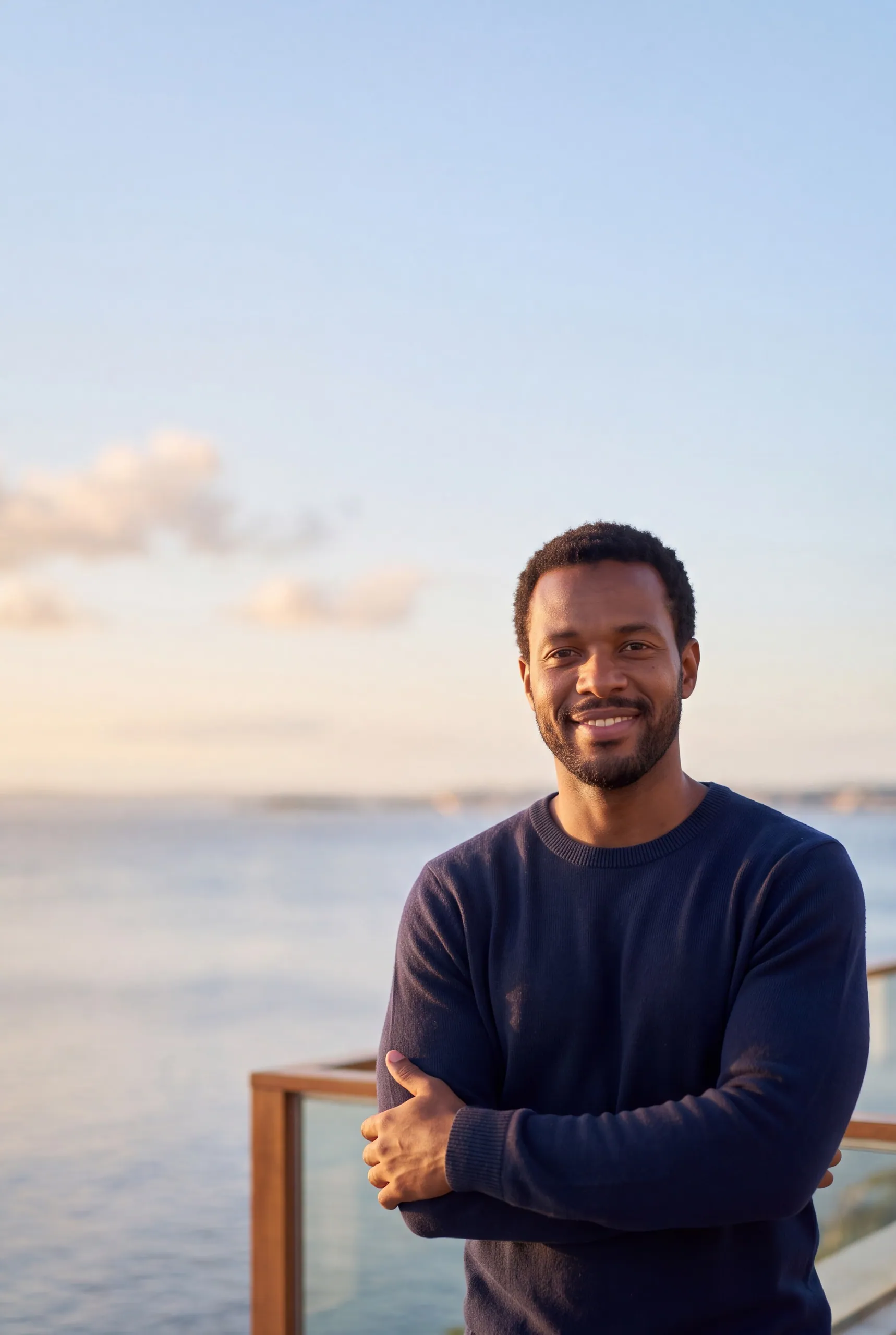 Homem sorrindo de braços cruzados em um terraço, com o mar ao fundo